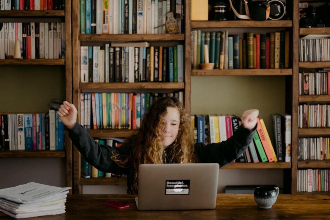a girl sitting in front of shelves of books, with an open laptop on the table in front of her raises her arms in triumph